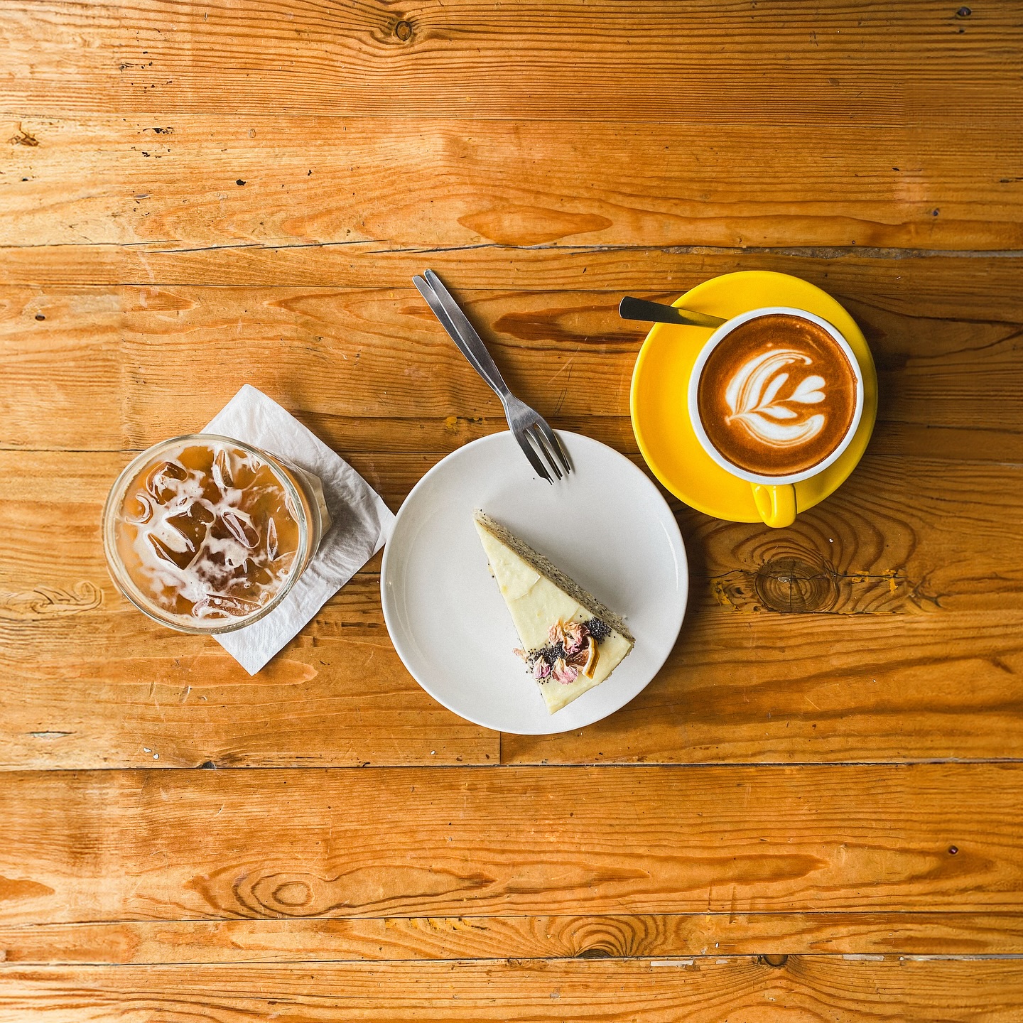 A flatlay on warm timber — an iced coffee, a slice of cake and a piccolo latte in a yellow cup